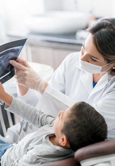 Una dentista con bata blanca y mascarilla mostrando una radiografía dental a un niño sentado en un sillón dental.